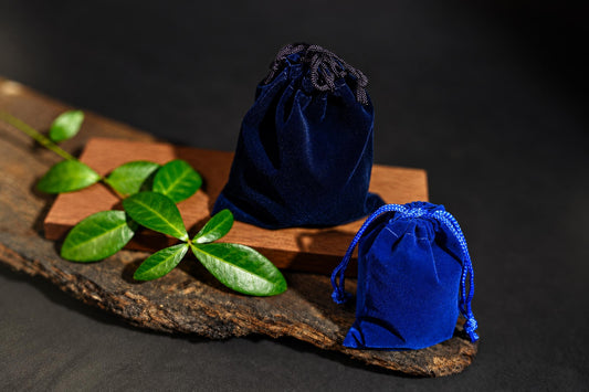 Two blue velvet drawstring pouches sit on a wooden surface with green leaves nearby.