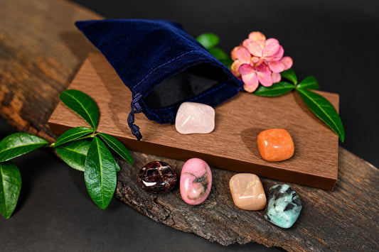 A blue velvet pouch with a drawstring sits on wood, spilling out six polished stones in pink, orange, red, and green.