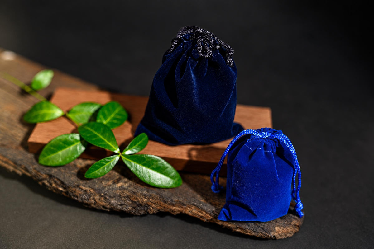 Two blue velvet drawstring pouches sit on a wooden surface with green leaves nearby.
