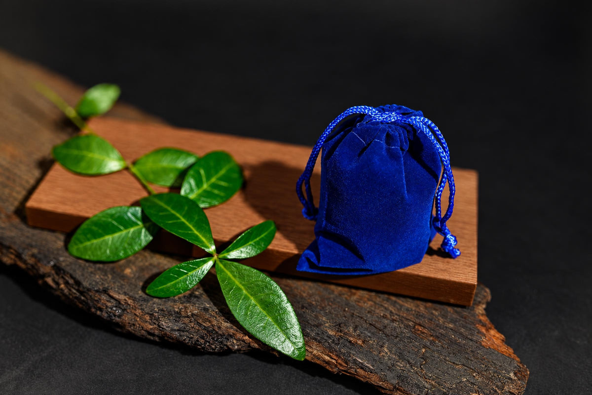 A blue velvet pouch with a drawstring sits on a wooden block next to green leaves on a dark surface.