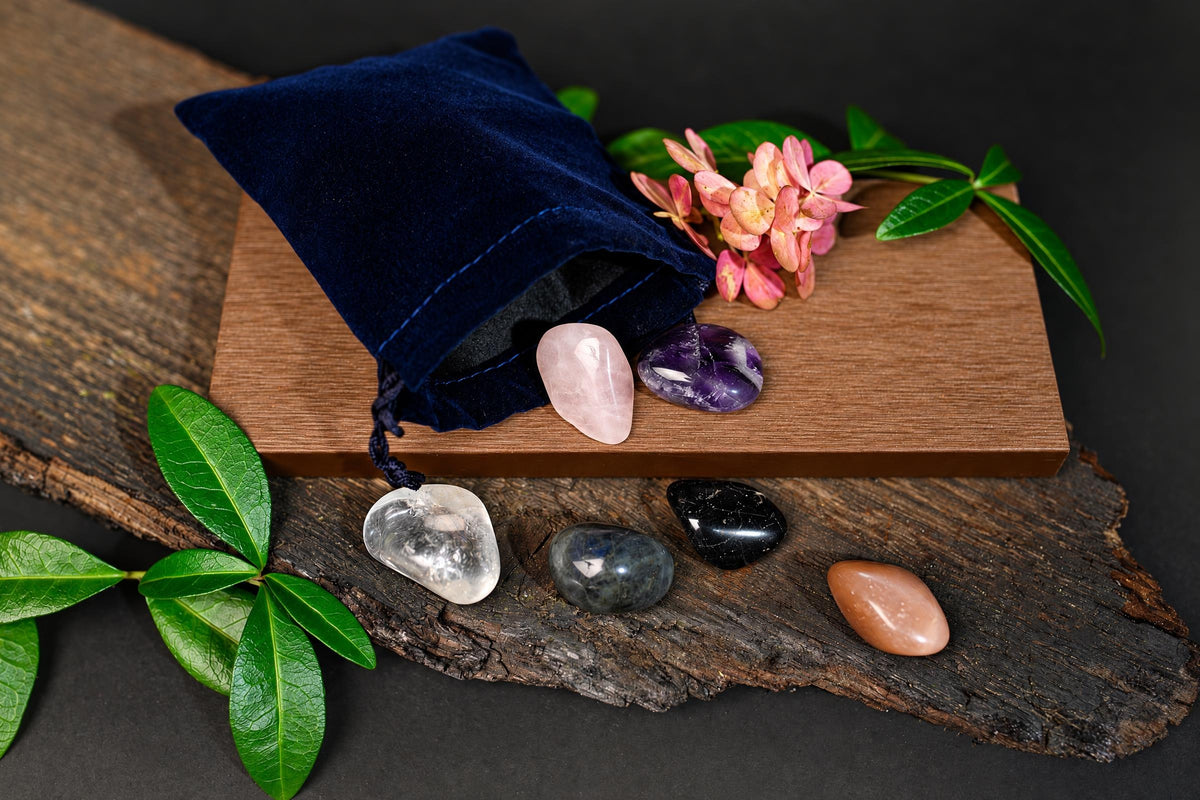 A dark blue velvet pouch sits on a wooden board with pink flowers, next to six polished stones in various colours like clear, pink, purple, black and brown.