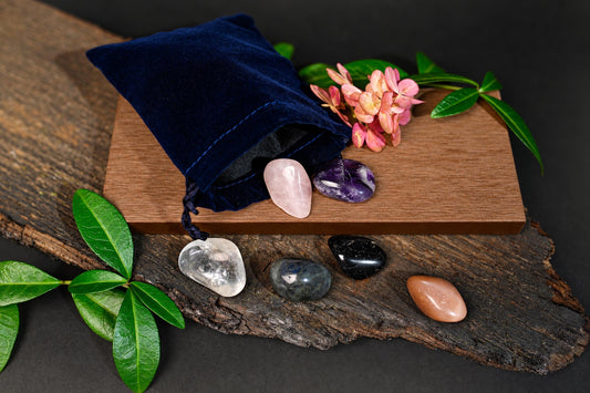A dark blue velvet pouch sits on a wooden board with pink flowers, next to six polished stones in various colours like clear, pink, purple, black and brown.