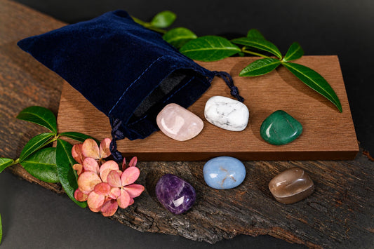 A dark blue velvet pouch with a drawstring sits on wood next to six polished stones in pink, white, green, purple, blue and brown.