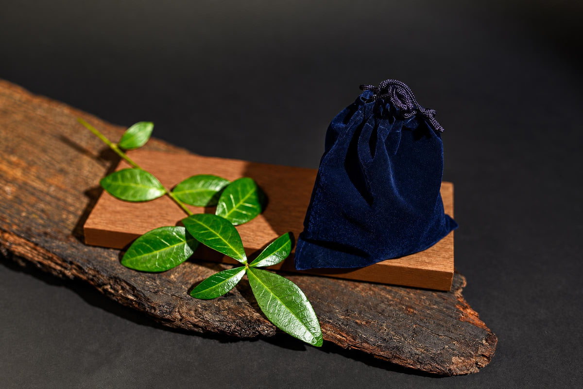 A dark blue velvet pouch with a drawstring sits on a wooden block, next to green leaves on a rustic wood slab.