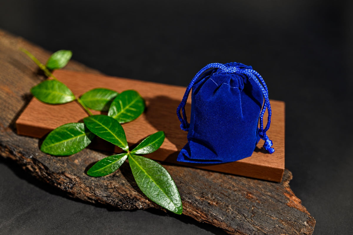 A blue velvet pouch with a drawstring sits on a wooden block next to green leaves.