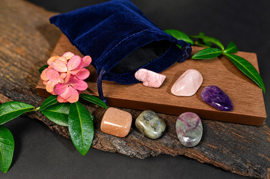 A blue velvet pouch sits on a wooden board with six polished stones in pink, purple, orange, and green hues, surrounded by leaves and a flower.
