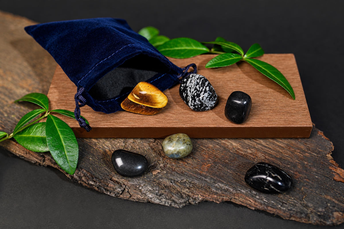 An open navy-blue pouch and polished black, brown and patterned stones rest on a wooden surface among green leaves.