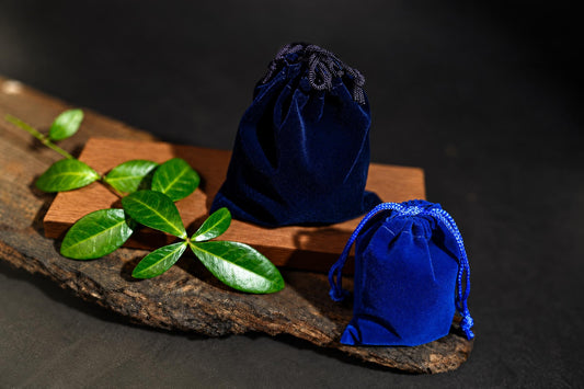 Navy-blue and bright blue drawstring pouches rest on a wooden surface beside green leaves against a dark background.