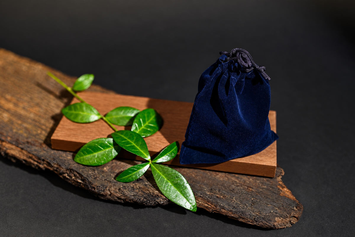 A navy-blue drawstring pouch stands on a wooden surface beside green leaves against a dark background.