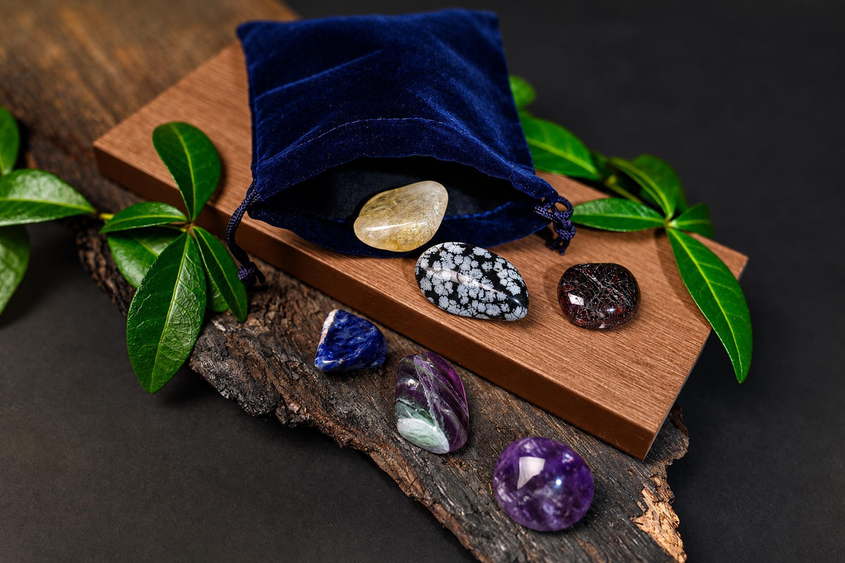 A blue velvet pouch with a yellow stone spills onto a wooden board and dark bark, surrounded by green leaves and other polished stones.