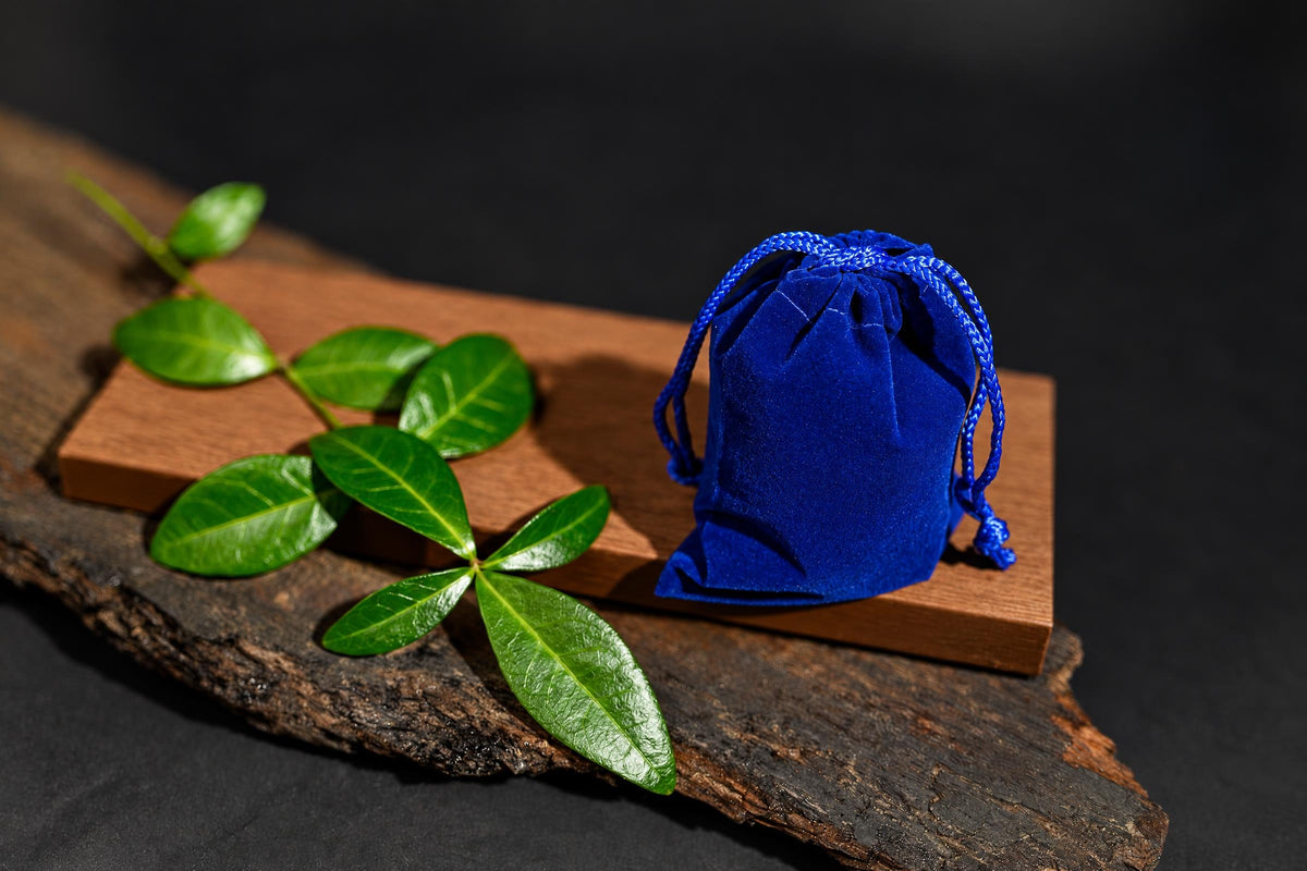 A blue velvet drawstring pouch sits on a wooden block with green leaves, all on a dark surface.