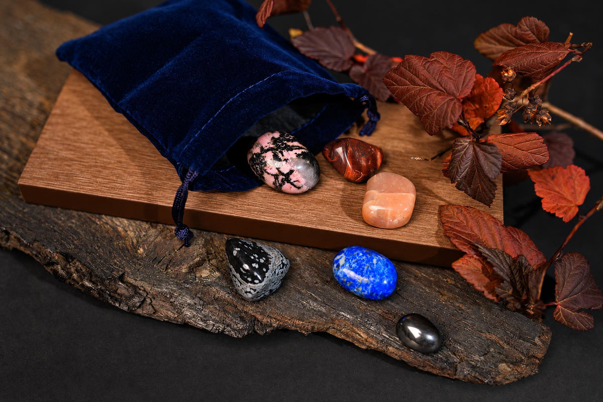 A blue velvet pouch on a wooden board with polished stones and dried leaves nearby.