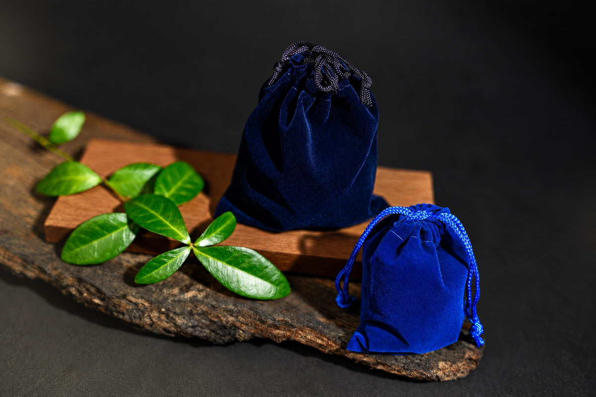 Two blue velvet drawstring pouches on a wooden surface with green leaves, one larger and one smaller.