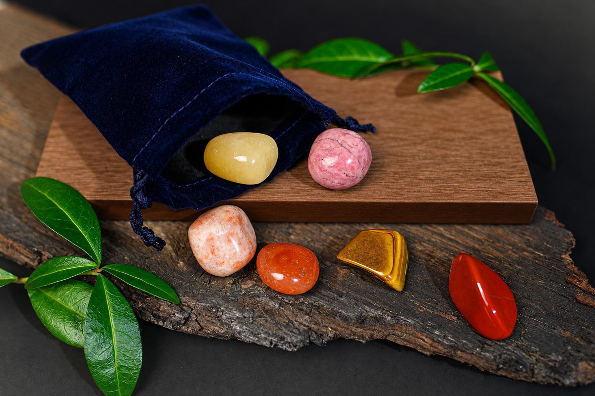 A dark blue pouch with yellow, pink, orange and red polished stones arranged on wood beside green leaves.