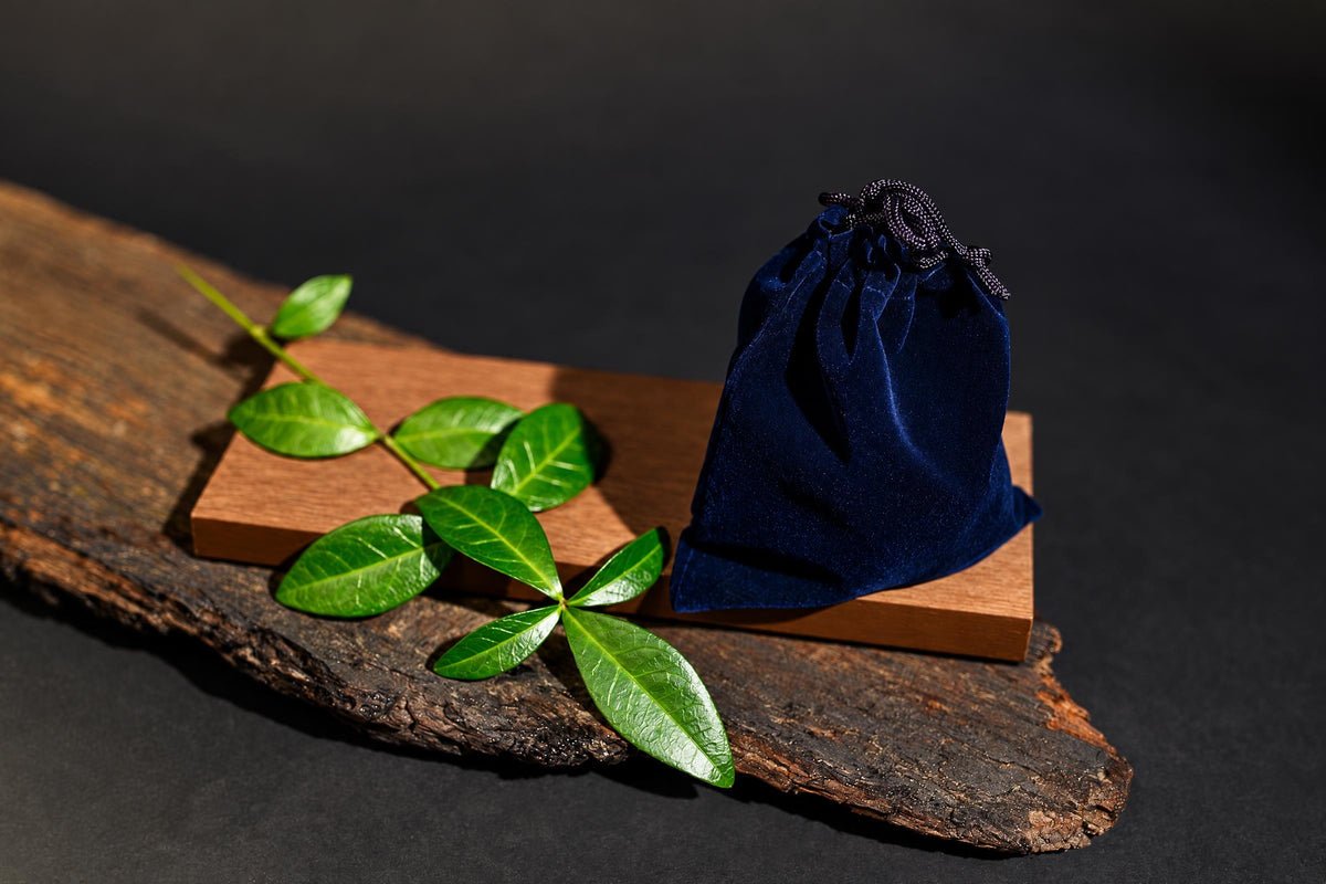 A dark blue velvet pouch with a black drawstring sits on a wooden block next to green leaves on a rustic wood slab.