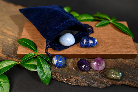 A blue velvet pouch on a wooden surface holds polished stones in shades of blue, white, purple, and green, surrounded by leaves.