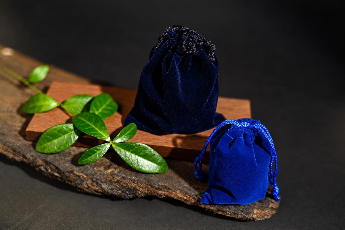 Two blue velvet drawstring pouches sit on a wooden surface with green leaves, against a dark background.
