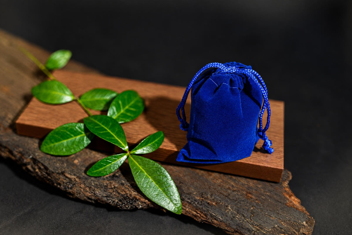 A blue velvet pouch with a drawstring sits on a wooden board next to green leaves.