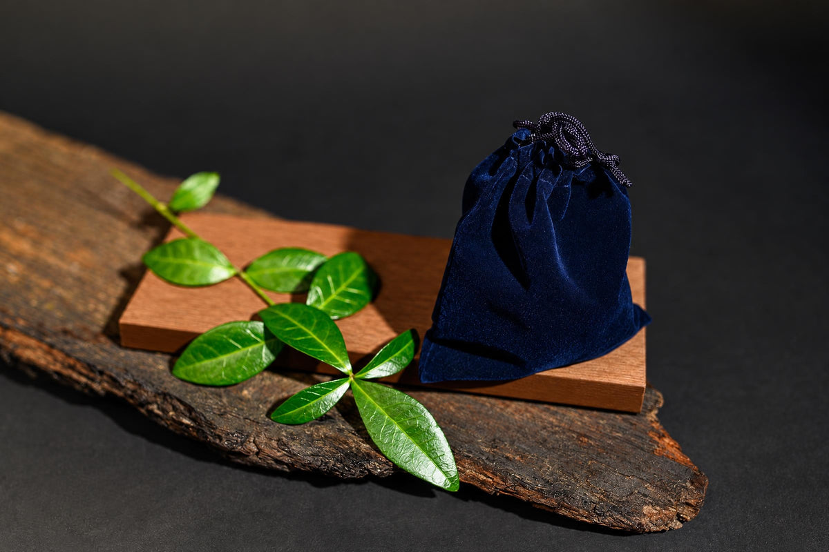 A dark blue velvet pouch with a drawstring sits on a wooden block, next to green leaves on a rustic wood slab.