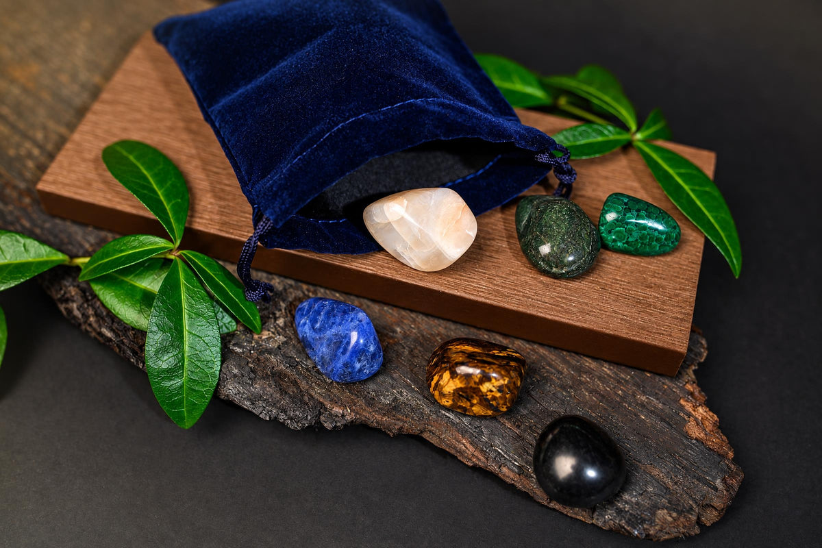 An open dark blue pouch and polished stones in varied colours sit on wooden surfaces framed by green leaves.