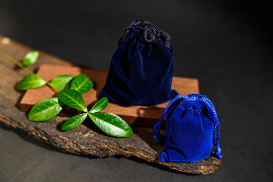 Two blue velvet drawstring pouches sit on a wooden surface with green leaves nearby.