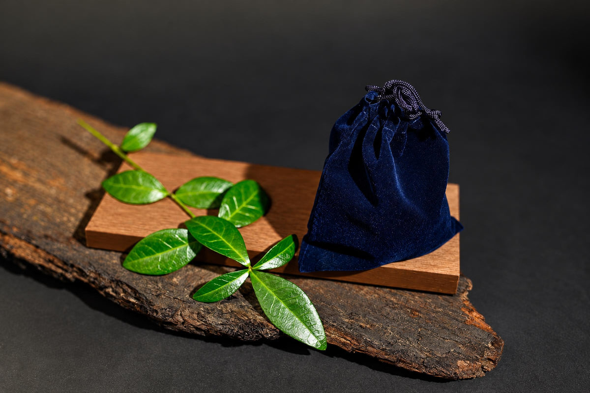 A dark blue velvet pouch with a drawstring sits on a wooden block next to green leaves on a rustic wood slab.