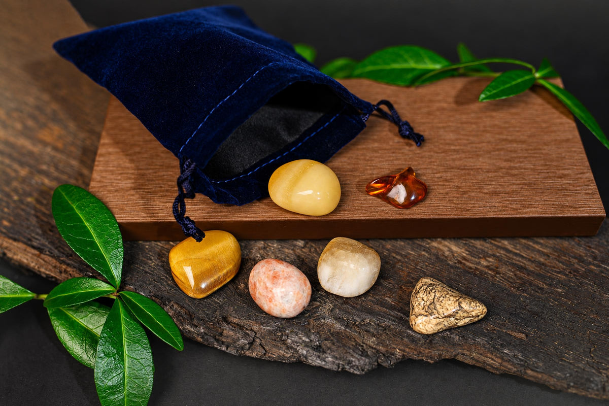 A blue velvet pouch with drawstrings sits on a wooden board, surrounded by six polished stones in yellow, orange, white, and patterned brown.