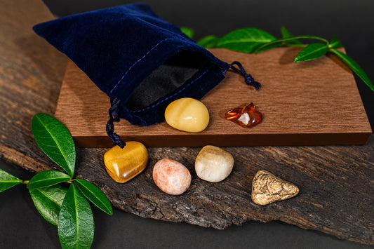 A blue velvet pouch with drawstrings sits on a wooden board, surrounded by six polished stones in yellow, orange, white, and patterned brown.