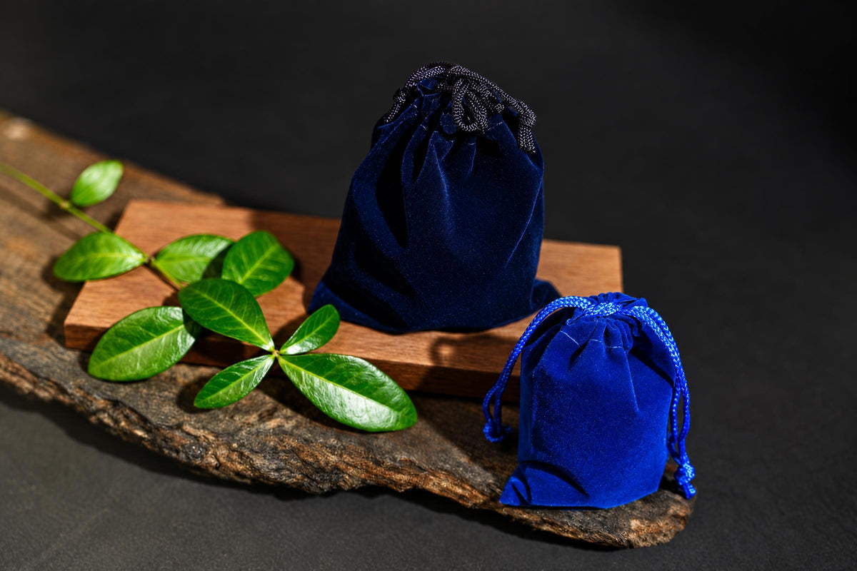 Two blue velvet drawstring pouches on a wooden surface with green leaves, one larger and one smaller.