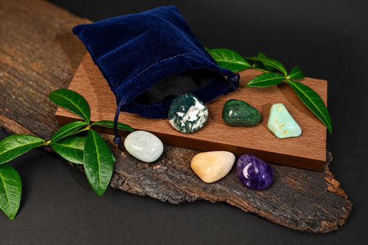 A blue velvet pouch sits on a wooden board with six polished stones in green, white, purple, and beige, arranged on wood and dark surface.