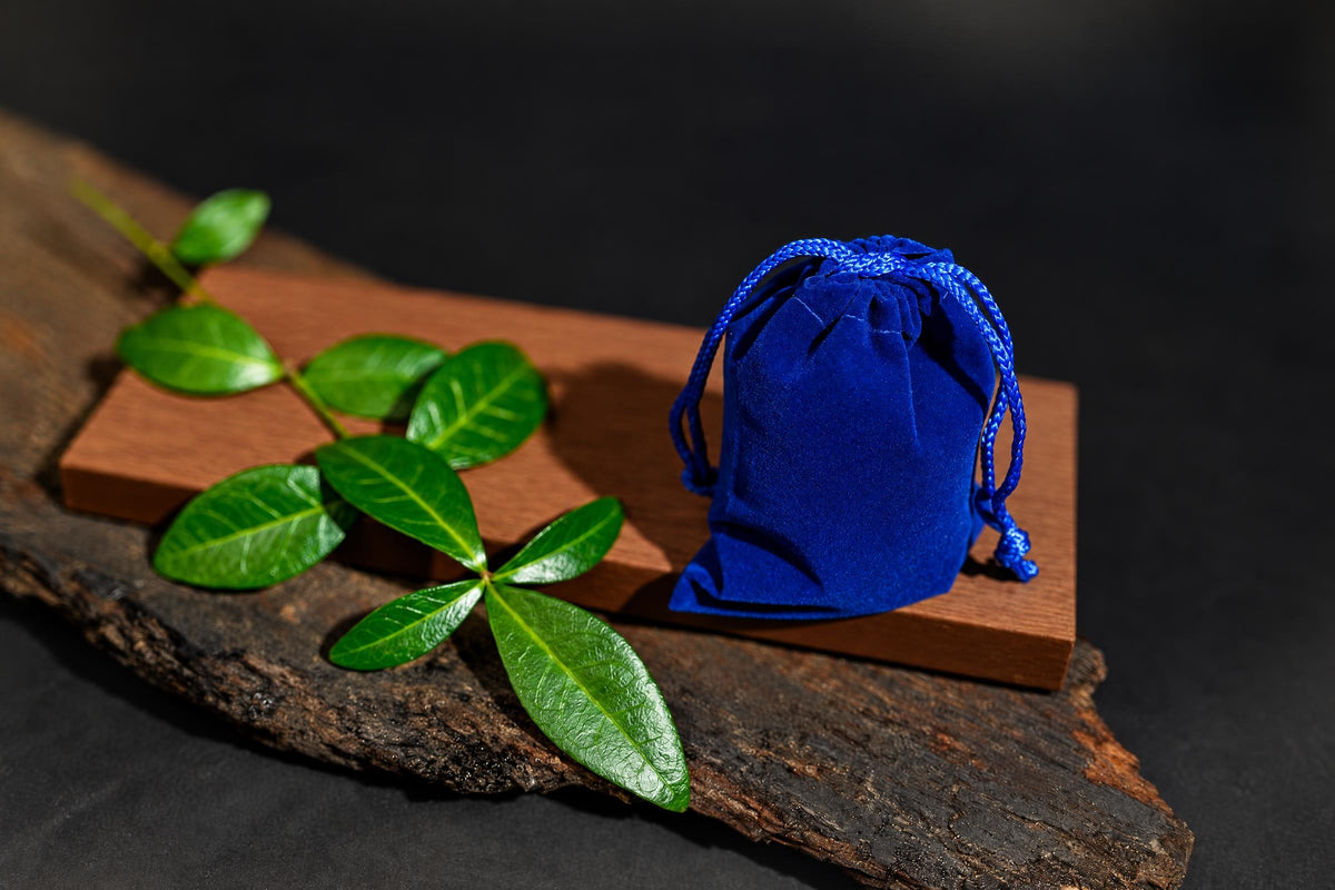 A blue velvet drawstring pouch sits on a wooden block with green leaves, all on a dark surface.