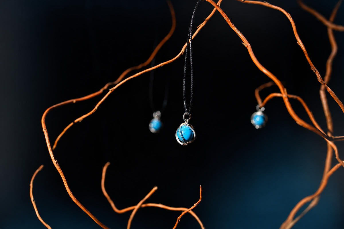 A bright blue spherical pendant in a silver-tone wrap hangs from a black cord among brown branches on a dark background.