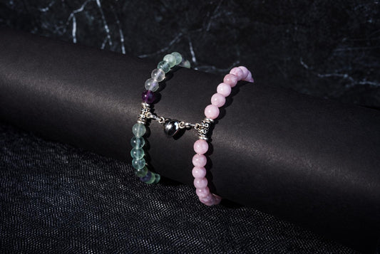 Two beaded bracelets on a dark cylindrical form: one with green and purple beads, the other pink, joined by a silver heart charm.