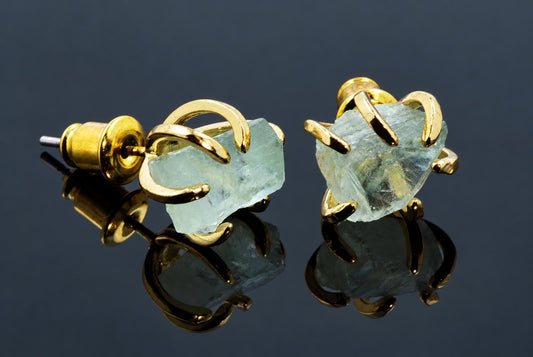 A pair of gold-toned stud earrings with curved metal claws holding pale green, rough-cut fluorite stones on a dark reflective surface.