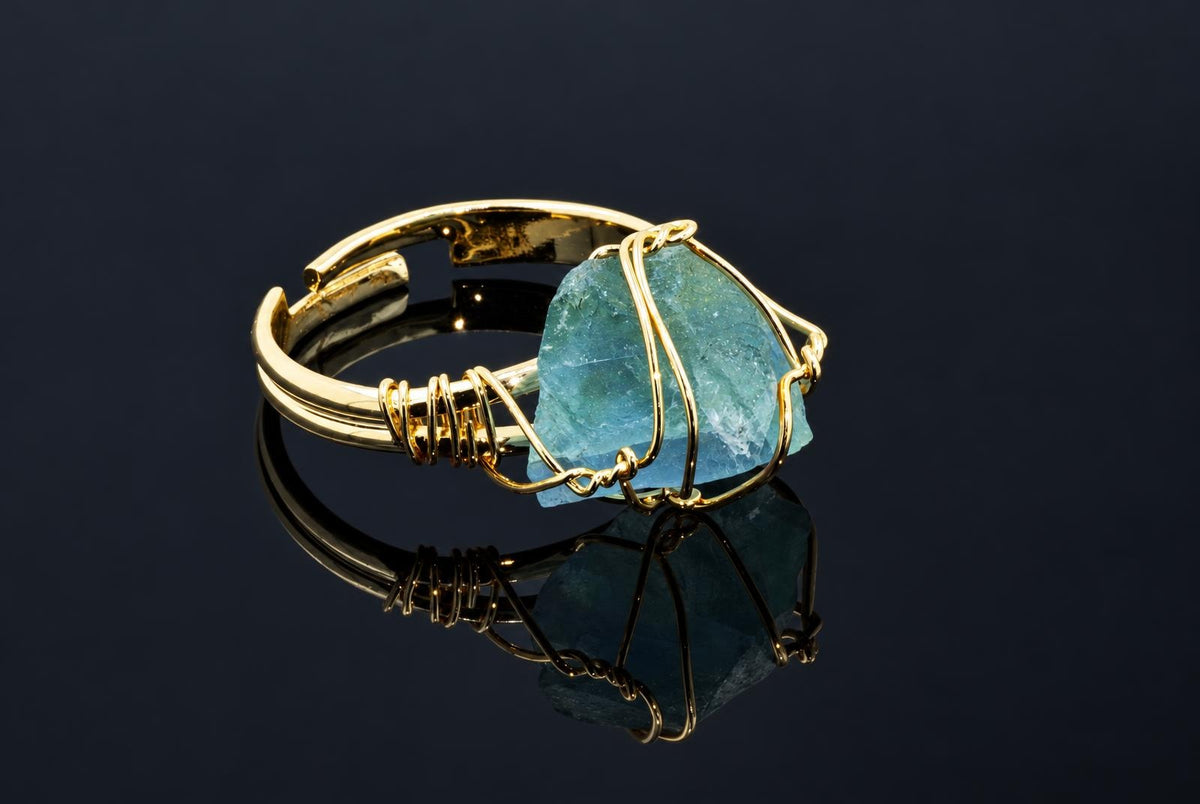 A gold-coloured ring holds a rough blue-green crystal wrapped in thin wire on a dark reflective surface.