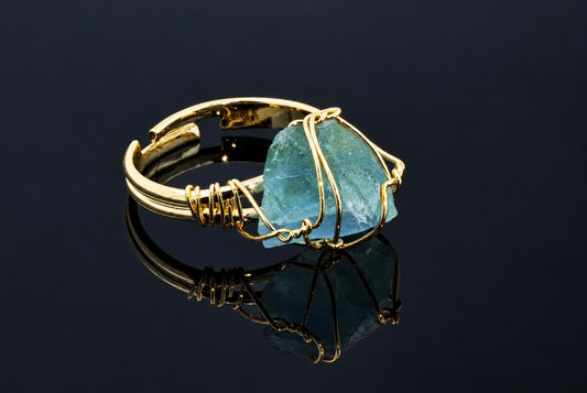 A gold-coloured ring holds a rough blue-green crystal wrapped in thin wire on a dark reflective surface.