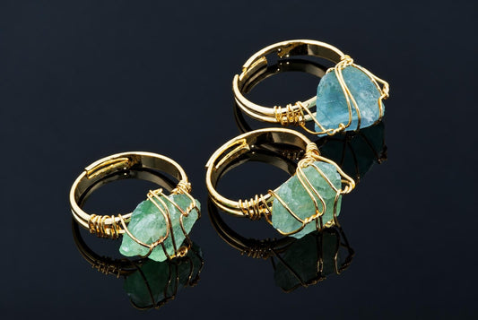 Gold-coloured rings hold rough green and blue crystals wrapped in thin wire on a dark reflective surface.