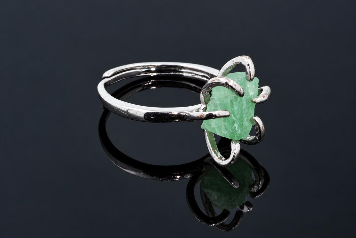 A silver-tone ring holds a rough pale green crystal in curved arms on a dark reflective surface.