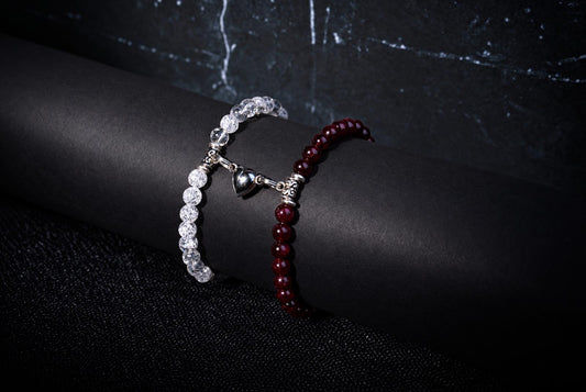 Two beaded bracelets, one clear and one red, with a silver heart charm, rest on a dark cylindrical surface.