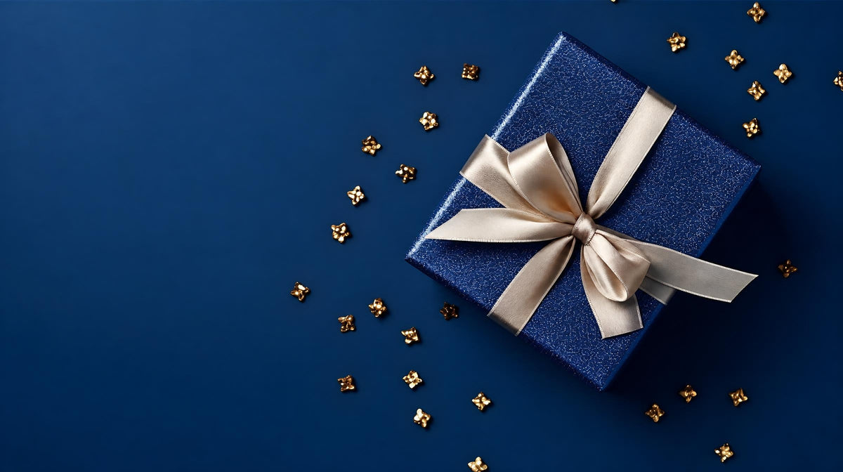 A blue gift box tied with a pale bow lies on a dark blue background scattered with small gold-coloured decorations.