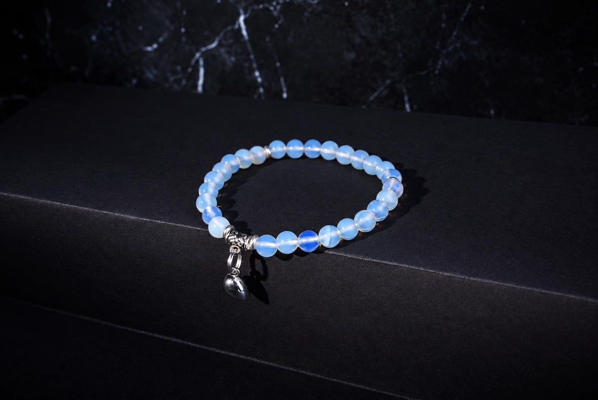 Glass opal bracelet with translucent pale blue round beads, ornate silver spacers and a hanging heart charm on a dark background.
