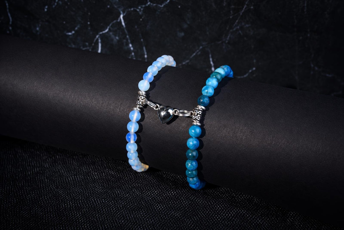 Two beaded bracelets with blue and white beads, silver heart charm, on a dark cylindrical surface.
