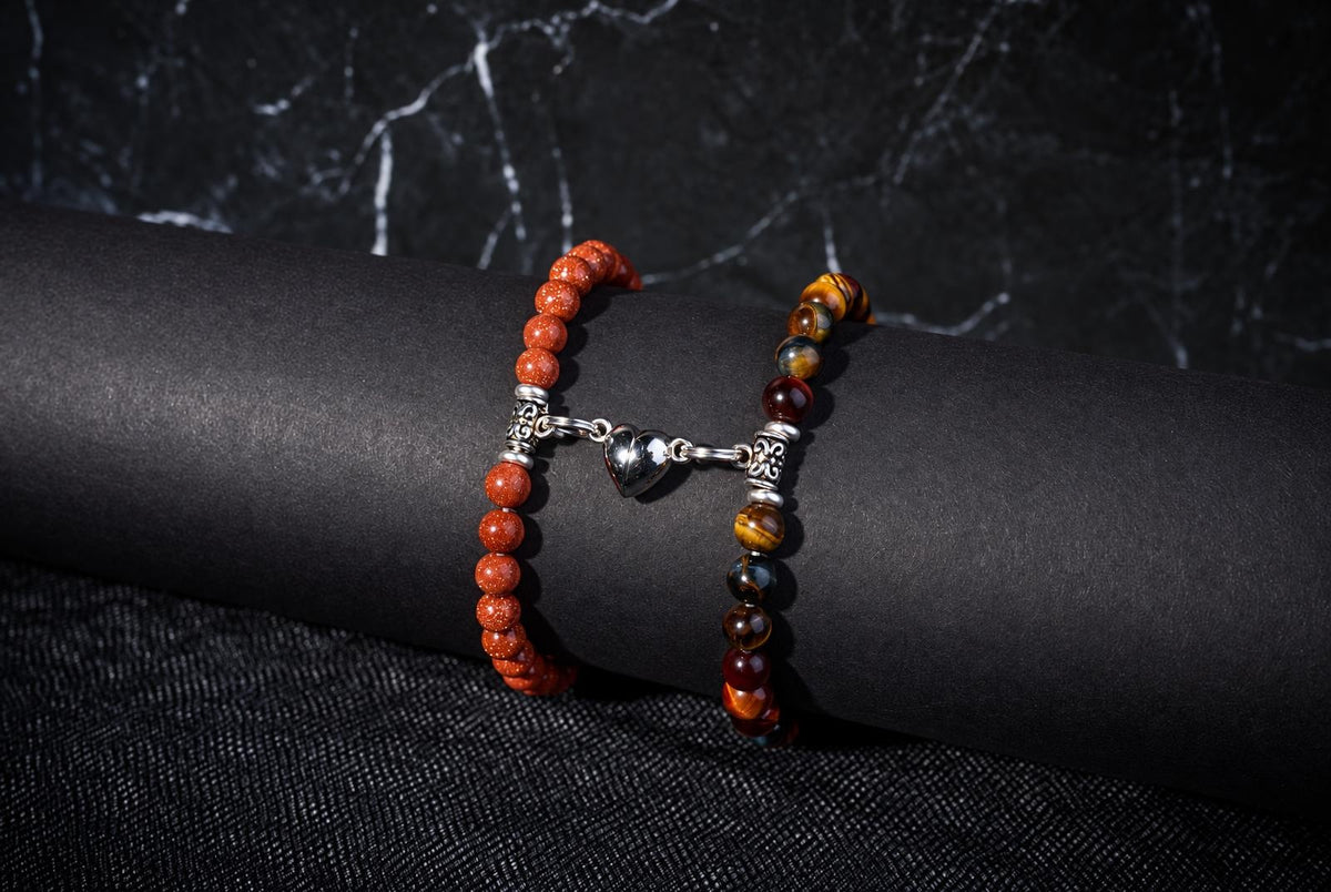Two beaded bracelets on a dark surface, connected by a silver heart charm. One bracelet has orange beads; the other has multicolored brown and yellow beads.