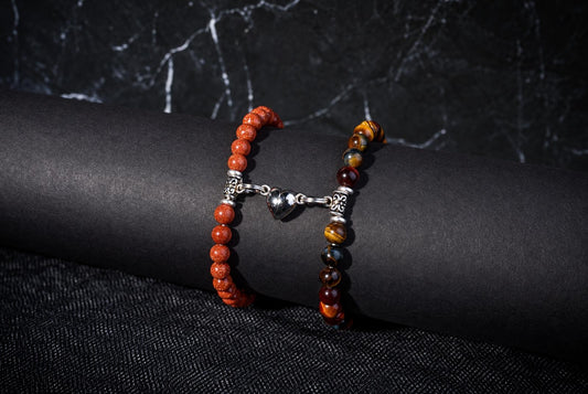Two beaded bracelets on a dark surface, connected by a silver heart charm. One bracelet has orange beads; the other has multicolored brown and yellow beads.