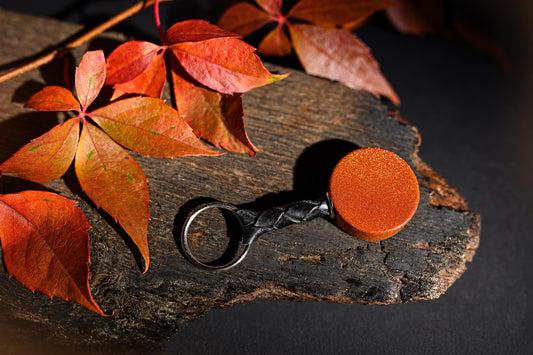 A keychain with a sparkling orange round disc, black braided strap and silver-tone ring rests on wood beside red-orange leaves.