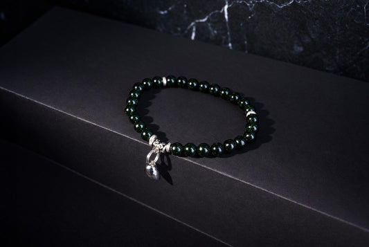A bracelet with dark green beads and silver accents, including a small bell charm, rests on a dark surface.