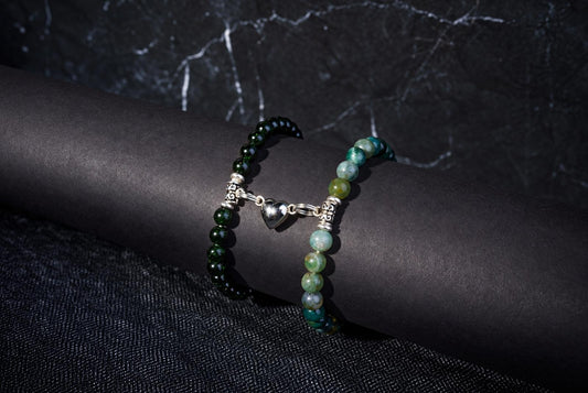 Two beaded bracelets with green and blue-green beads, silver spacers, and a heart charm on a dark cylindrical form.