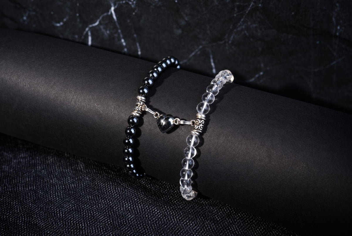 Two bracelets on a dark surface: one with black beads and a silver heart charm, the other with clear beads.