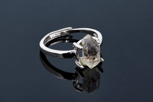 A silver-tone ring holds an elongated clear crystal with dark inclusions on a dark reflective surface.