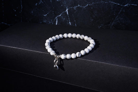 Howlite bracelet with round white and grey veined beads, ornate silver spacers and a hanging heart charm on a dark background.